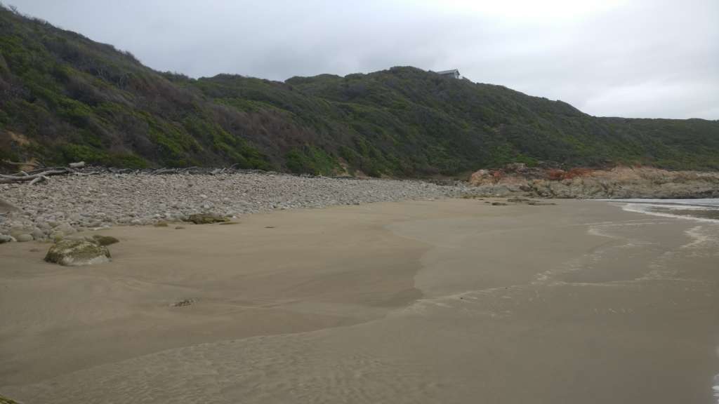 Une plage de sable au premier plan, avec l'océan derrière le photographe. Au milieu se trouve une partie rocheuse de la plage, avec une colline au-delà. A l'horizon au sommet de la colline se trouve la maison Fonteintjies.